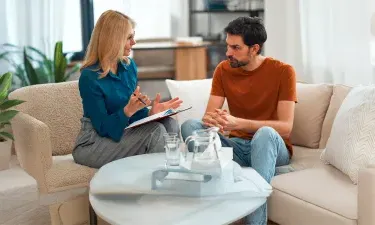 A counselor, wearing a blue blouse and gray skirt, is engaged in a conversation with a male client in a casual therapy session.